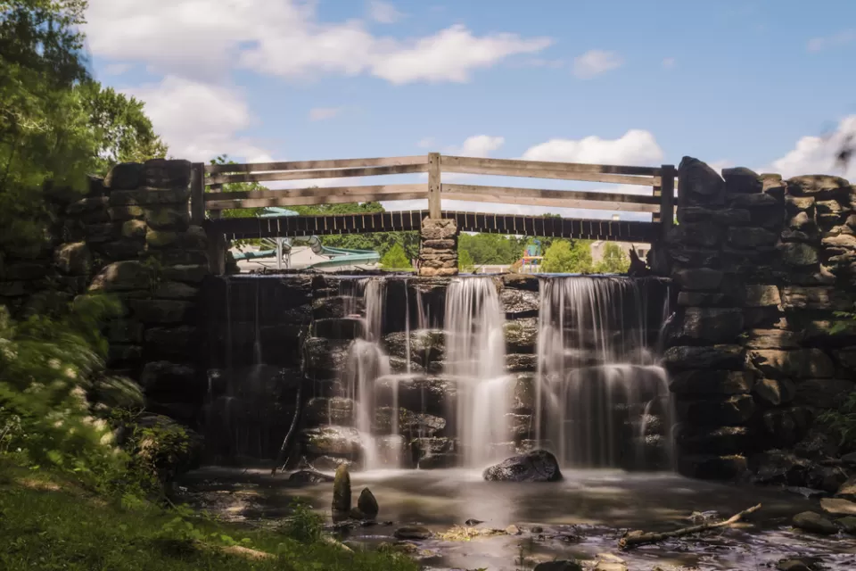 A footbrinde crosses over a small cascade of flowing water on a sunny day in Yonkers, New York.
